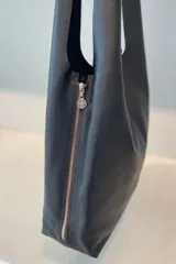 Bolso tipo tote de gran capacidad, confeccionado artesanalmente en cuero vacuno color marrón. Presenta un diseño minimalista con costura central vertical, fuelle lateral ajustable con cierre y placa metálica con logo en la parte inferior.