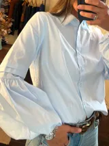 Camisa de algodón color celeste, con cuello clásico, cierre frontal con botones y mangas largas abullonadas con detalle de pliegues en el antebrazo.