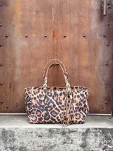 Cartera tipo tote con estampado animal print de leopardo. Cuenta con doble asa de hombro ajustable con hebillas metálicas y detalle de flecos colgantes en uno de los laterales.