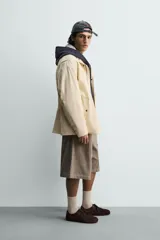 Parka de tejido técnico color beige claro, corte relajado (relaxed fit), con cuello subido, cierre frontal oculto por solapa con botones a presión y bolsillos de solapa en cadera.