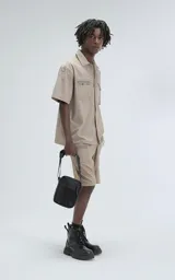 Camisa de manga corta color beige, estilo técnico, confeccionada en microfibra liviana. Cuenta con doble bolsillo frontal, uno con detalle de malla y otro con cierre, y broches en lugar de botones.