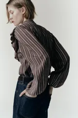 Camisa de manga larga con cuello subido, cierre frontal con botones y detalle de volantes en el pecho. Presenta un diseño a rayas verticales y puños elásticos.