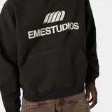 Hoodie negro con capucha, bolsillo tipo canguro y estampado frontal blanco con el logo de "EMESTUDIOS".