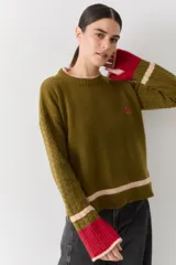 Buzo de lana de corte regular en color verde oliva, con mangas tejidas en ochos y puños acanalados en color rojo con detalle en rosa. Presenta un bordado de pelota de tenis en el pecho y aberturas para los pulgares en los puños.