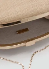 Bolso tipo clutch de diseño ovalado y rígido, revestido en rafia color tostado, con cierre metálico a presión en la parte superior y detalles metálicos.