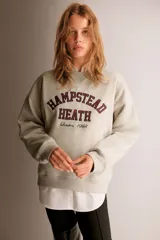 Buzo oversized color beige con interior cepillado suave. Presenta hombros caídos y un diseño de texto en el pecho que dice "HAMPSTEAD HEATH London, 1998" en rojo y negro. Tiene ribete acanalado en cuello, puños y bajo.