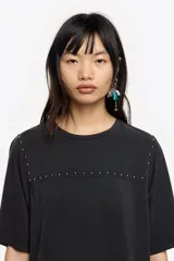 Camiseta de manga corta y escote redondo, color beige claro, con corte boxy. Presenta una hilera de tachuelas metálicas negras decorativas en la parte superior del pecho y en las costuras de los hombros.