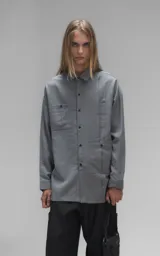 Camisa de manga larga en tejido twill color gris melange, con corte oversize. Presenta cuello clásico, cierre frontal con botones y tres bolsillos delanteros, uno de ellos oculto.