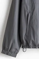 Campera rompevientos de nylon color gris oscuro, con forro de malla. Presenta cierre frontal con protección para el mentón, capucha ajustable con cordón, bolsillos laterales y un bolsillo interior, todos con cierre. Los puños y el bajo tienen elástico revestido.