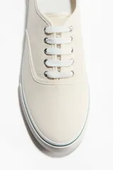 Calzado deportivo de lona de algodón color crudo, con cordones y suela de goma blanca con una línea verde.
