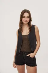 Chaleco corto de denim negro con escote en V y diseño sin mangas. Presenta terminación lavada y detalles laterales con entramado y tachas metálicas que aportan un toque distintivo.