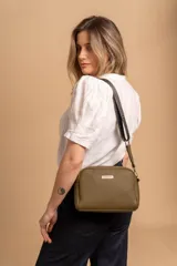 Bandolera de diseño compacto con textura granulada, color verde oliva. Cuenta con una correa ajustable de tela negra con el logo de la marca en relieve y placa metálica frontal con el nombre de la marca.