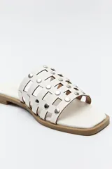 Sandalia baja de diseño entrelazado con tiras negras y apliques metálicos circulares. Presenta una suela plana de color marrón y puntera cuadrada.