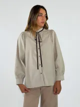 Camisa de corte holgado en color beige, con cuello alto estilo victoriano, detalle de volados en el cuello y lazo de cinta oscura para anudar. Presenta mangas largas abullonadas con puños ajustados y cierre frontal con botones.