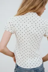 Top de popelina de color blanco con estampado de lunares negros, cuello mao con escote en pico y lazada, mangas cortas abullonadas y cierre frontal con botones.