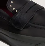 Championes Vans tipo mocasín de cuero negro, con diseño inspirado en el skate y estética clásica. Presenta detalles de tachuelas metálicas en la lengüeta, construcción vulcanizada y suela waffle de goma gris oscuro.