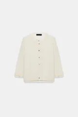 Chaqueta efecto doble de punto color beige con cuello redondo, manga por debajo del codo y cierre frontal con botones. Detalle de cuello y bajo de camisa interior asomados.
