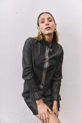 Camisa de jean gris oscuro con cuello mao, cierre frontal con botones metálicos y una solapa en contraste de color gris claro. Presenta mangas largas con puños elásticos fruncidos y volados en los extremos.