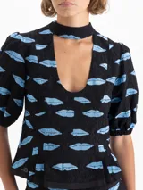 Top negro de jacquard con estampado de boquitas celestes. Presenta cuello con lazo, escote en V, mangas abullonadas con puños elásticos y un volado en el ruedo. Incluye recortes en el delantero y cierre trasero metálico.