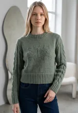 Sweater de punto color verde, con cuello redondo, diseño de trenzas en las mangas y detalle de relieve en el frente. Presenta un corte corto con puños y ruedo acanalados.