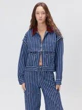 Campera de denim a rayas verticales con cuello de pana en color bordó. Presenta cierre frontal, mangas desmontables con cierre en los hombros, canesú en el pecho y bolsillos de parche cuadrados.