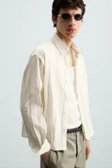 Camisa relaxed fit de manga larga con cuello solapa, confeccionada en tejido con mezcla de algodón. Presenta un estampado de rayas verticales en tonos beige, blanco y marrón. Incluye un bolsillo plastrón en el pecho, puños con botón y aberturas laterales en el bajo. Cierre frontal de botonadura.