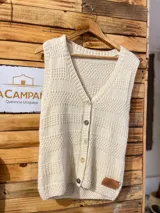 Chaleco tejido de lana merino 100%, color marfil, con escote en V, cierre frontal con botones y diseño texturado. Presenta hombros anchos y un pequeño aplique de cuero con grabado en el inferior.