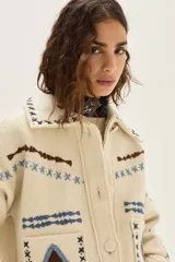 Campera de lana merino hervida en color crudo, con diseño de cuello camisero, cierre frontal de botones y bolsillos frontales. Presenta bordados artesanales con motivos geométricos en tonos marrón y azul en el frente, espalda y mangas.