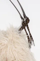 Bolso de hombro con estructura blanda, confeccionado en poliéster con acabado de pelo largo en color beige. Presenta asas de cuero marrón y detalles de cordones trenzados. Incluye cierre de cordón y bolsillo interior.