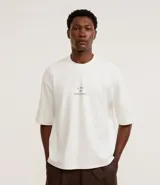 Remera boxy de algodón color crudo, con cuello redondo y mangas cortas. Estampa frontal con la frase "l'ami du contraire" y estampa trasera de figuras estilo art noveau.