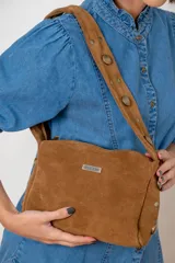 Cartera de hombro confeccionada en gamuza color marrón, con correa ancha decorada con tachas metálicas circulares. Presenta una placa metálica con el logo de la marca en el frente y cierre superior.
