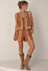 Conjunto de dos piezas color tostado con bordados florales en hilo blanco, compuesto por una blusa sin mangas con escote en V y cierre frontal con lazos, y un short tiro alto.