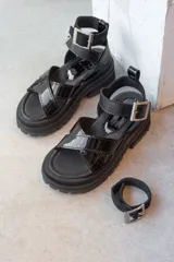 Sandalias negras de estilo chunky con tiras cruzadas en charol y base de goma dentada. Cuentan con una tira superior desmontable y cierre con hebilla metálica en el tobillo.