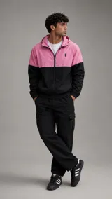 Conjunto deportivo compuesto por una campera con capucha y cierre frontal, de diseño bicolor en rosa y negro, y un pantalón cargo negro a juego. La campera presenta un pequeño logo bordado en el pecho.