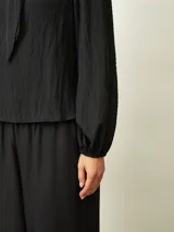 Blusa de textura arrugada con cuello alto y lazo frontal para anudar. Presenta mangas largas con puños elásticos y corte recto.
