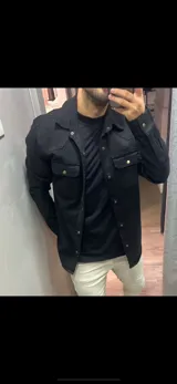 Sobrecamisa de gabardina negra con cierre frontal de botones metálicos a presión, bolsillos frontales con solapa y mangas largas.