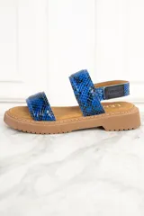 Sandalias con estampado de piel de serpiente en tonos azules, con dos tiras anchas sobre el empeine y una tira alrededor del tobillo. La suela es plana y de color marrón claro.