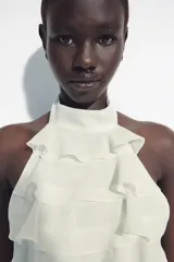 Top de cuello halter color blanco, con múltiples volantes en cascada en todo el frente y la espalda descubierta con cierre de botones y lazada en el cuello.