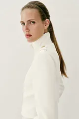 Chaqueta corta blanca de estilo militar, con cuello subido, manga larga y trabillas desflecadas en los hombros. Cuenta con cierre frontal cruzado decorado con botones metálicos dorados.