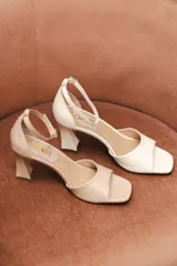 Sandalia de tacón color beige, con punta cuadrada abierta, talón cerrado y pulsera ajustable al tobillo con hebilla dorada. El tacón es de 6cm, con diseño geométrico que se afina en el centro.