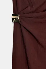 Vestido midi color bordó con escote asimétrico y manga sisa. Presenta frunces en el delantero con un aplique metálico dorado en forma de nudo.
