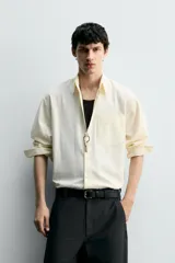 Camisa de algodón color crema o blanco roto, de corte regular fit, con cuello solapa, manga larga con puño abotonado y bolsillo plastrón en el pecho. La tela presenta una textura sutil de rayas verticales en relieve.