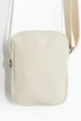 Bandolera de lona color crudo con correa ajustable. Tiene dos compartimentos exteriores, uno abierto y otro con cierre, y un compartimento interior abierto.