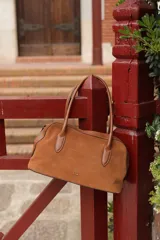 Cartera tipo bowling de cuero gamuza color tostado, con doble asa de mano en cuero liso, cierre superior y logo metálico de la marca en el frente.