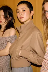 Chaqueta bomber de corte cropped confeccionada en material efecto piel color beige, con cuello subido, puños y cintura en rib elástico a tono, cierre frontal con cremallera y bolsillos laterales de vivo.