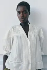 Camisa blanca de algodón con cuello redondo y manga por debajo del codo abullonada. Presenta detalles de bordados a tono y cierre frontal con botones.