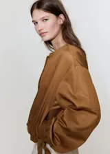 Chaqueta de diseño corto en tono tostado, confeccionada con mezcla de lyocell, algodón y lino. Presenta cuello redondo, mangas largas abullonadas, elásticos en los laterales y cierre frontal de botonadura visible.