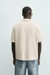 Camisa de manga corta y calce relajado en color beige, con cuello bowling y cierre frontal de botones. Presenta un diseño bordado en la parte inferior con motivos de paisaje tropical que incluyen palmeras, sol y aves.