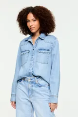 Camisa de denim liviano de color azul claro, con inspiración western, cuello clásico, cierre frontal con broches metálicos y bolsillos delanteros con solapa. Presenta un calce relajado y mangas largas con puños abotonados.