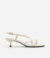 Sandalia femenina confeccionada en material sintético color crema, con punta redonda, tiras finas asimétricas, dedera con detalle bold y hebilla lateral. Prenda con salto mediano fino.